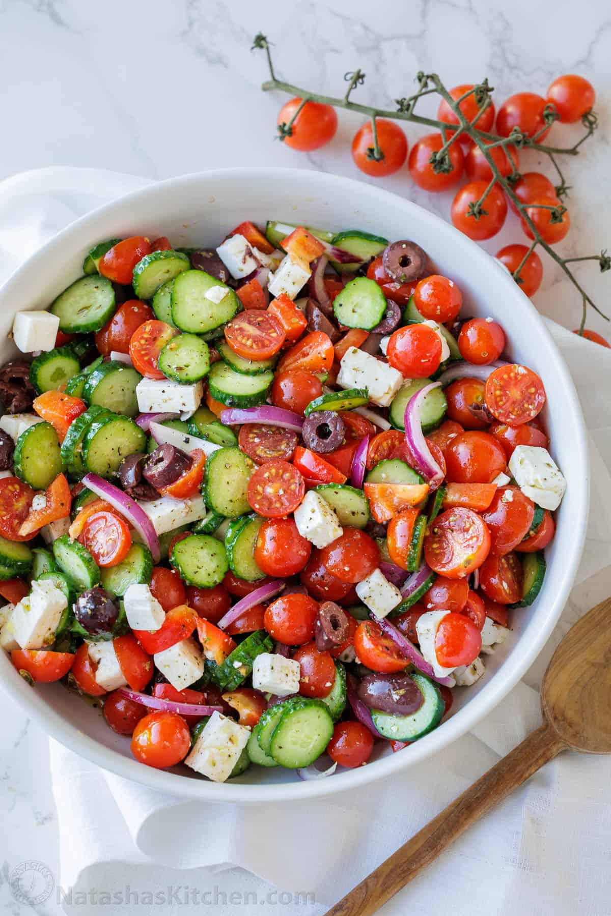 Classic Greek Salad recipe in a white bowl with cherry tomatoes and a wooden spoon on the side