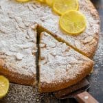 A slice of almond cake removed from the whole with almond slices and confectioners sugar on top