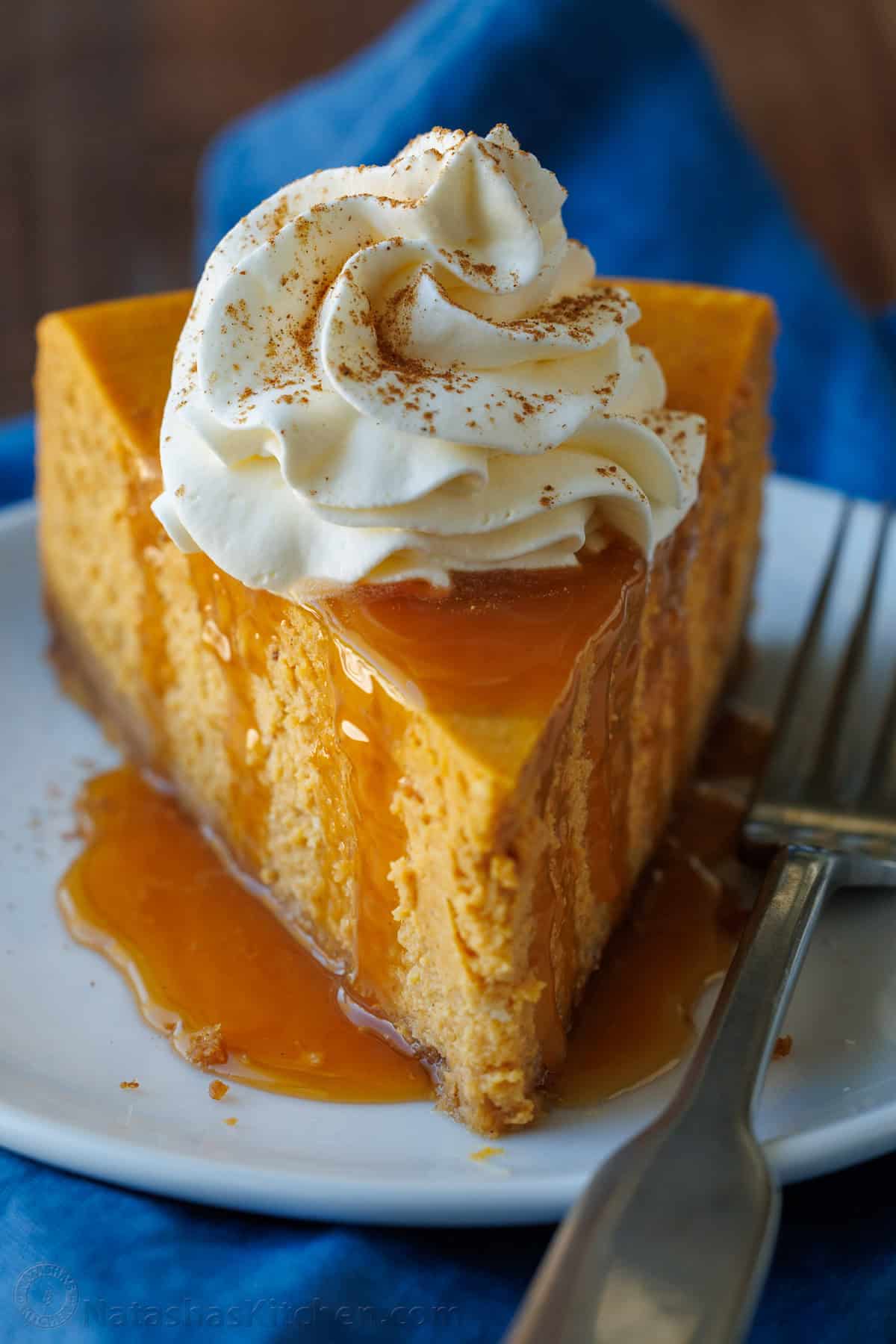 My pumpkin cheesecake recipe is so easy to make and is better than any professional baker! Save money and make your Holiday dessert at home! A slice of pumpkin cheesecake with whipped cream on a white plate with a blue napkin.