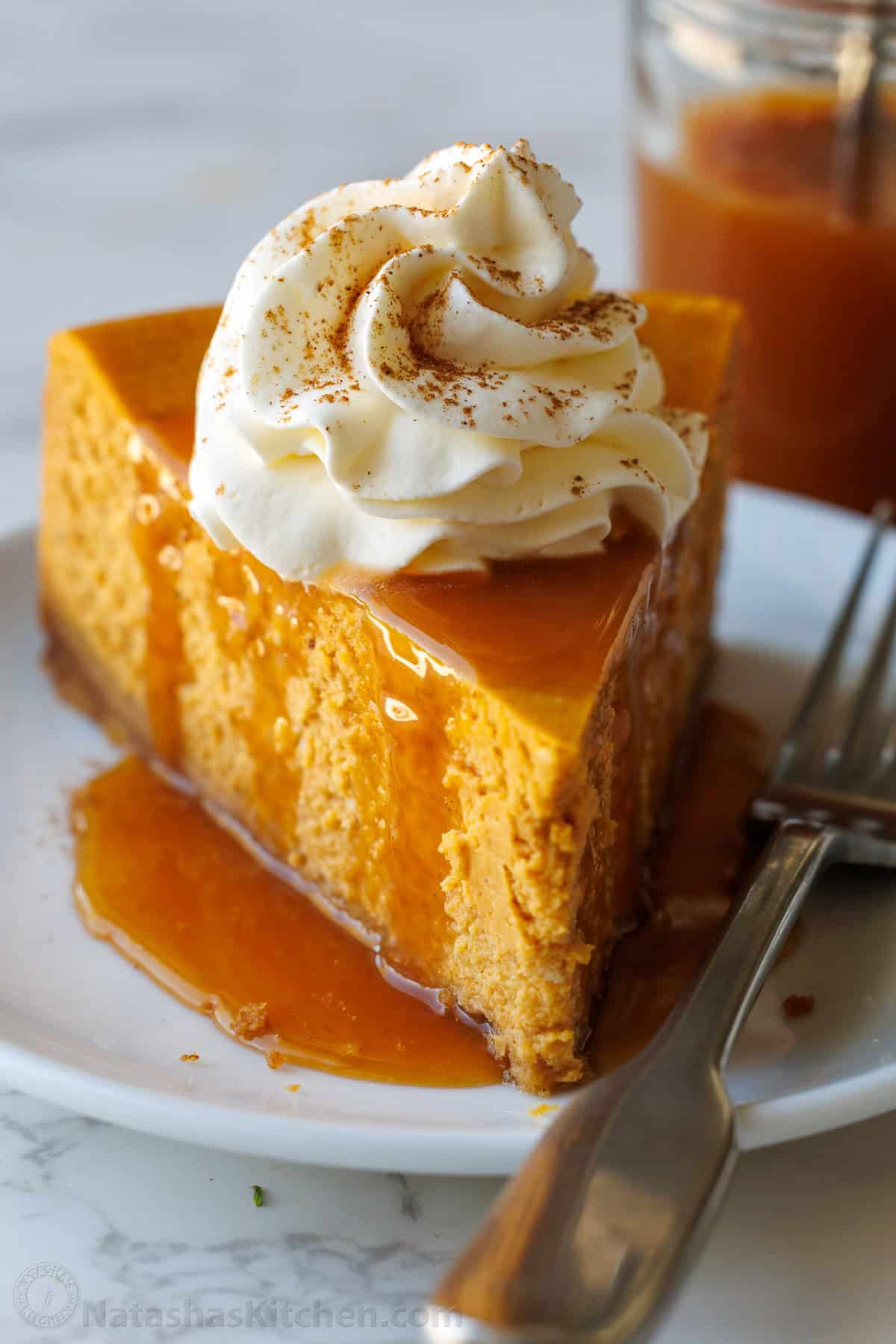 My pumpkin cheesecake recipe is so easy to make and is better than any professional baker! Save money and make your Holiday dessert at home! Pumpkin cheesecake garnished with whipped cream and a dusting of cinnamon on a plate with a fork