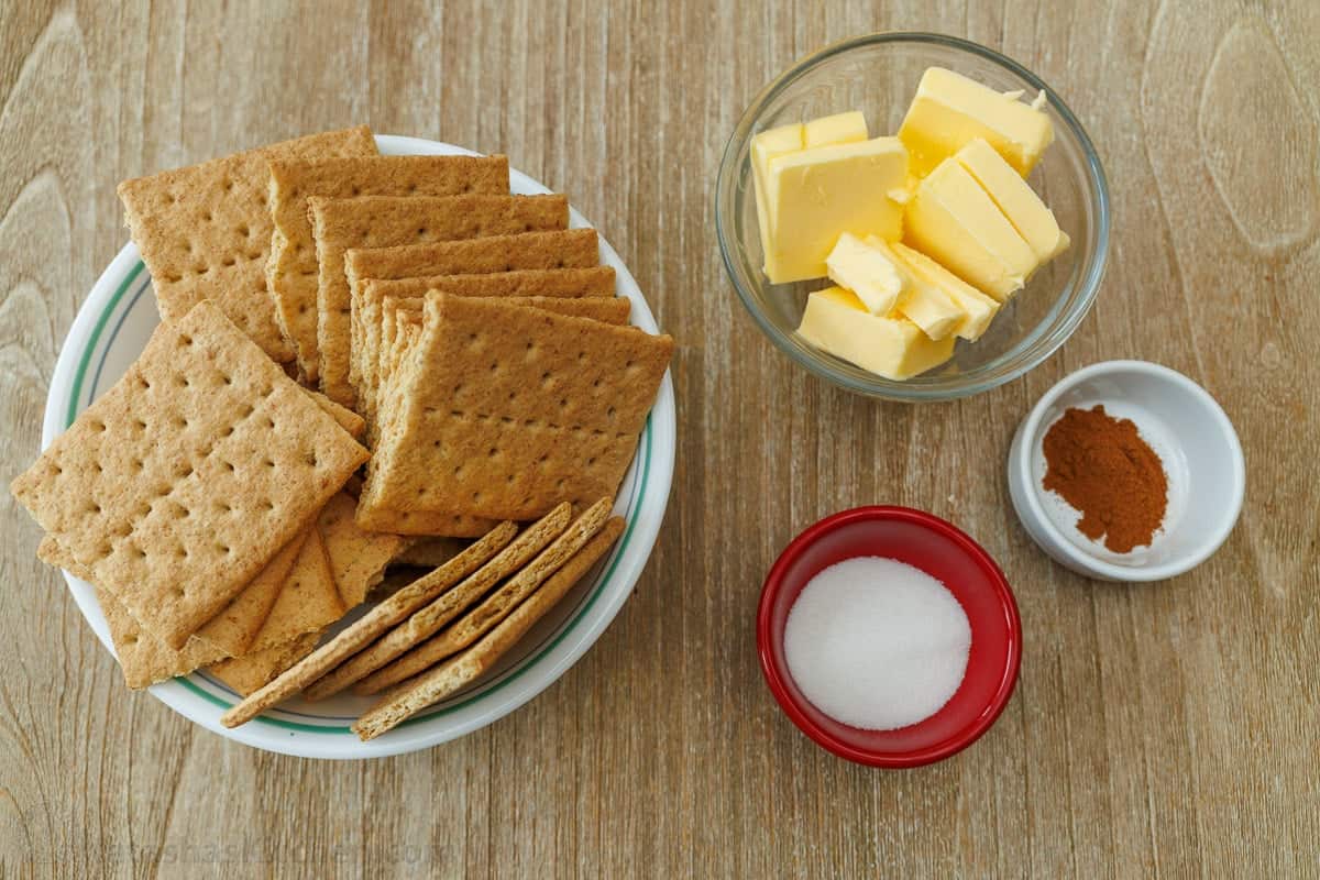 My pumpkin cheesecake recipe is so easy to make and is better than any professional baker! Save money and make your Holiday dessert at home! ingredients for making graham cracker crust, including graham crackers, butter, cinnamon and sugar in bowls on a table.