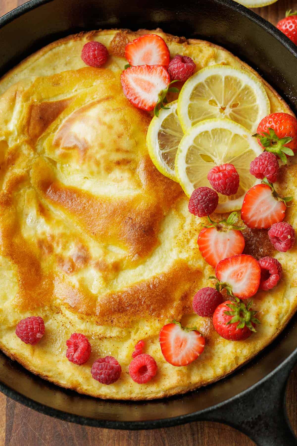 Easy Sourdough Dutch Baby pancake recipe using sourdough discard. Blend or whisk the batter, and it cooks up puffy and golden in the oven. Close up of a German Pancake in a cast iron pan with crispy brown edges and garnished with berries and sliced strawberries