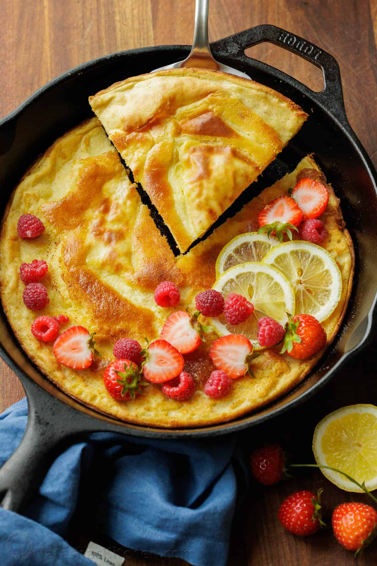 Easy Sourdough Dutch Baby pancake recipe using sourdough discard. Blend or whisk the batter, and it cooks up puffy and golden in the oven. Sourdough Discard Dutch Baby pancake slice lifted out of a cast iron pan garnished with berries and lemon slices.