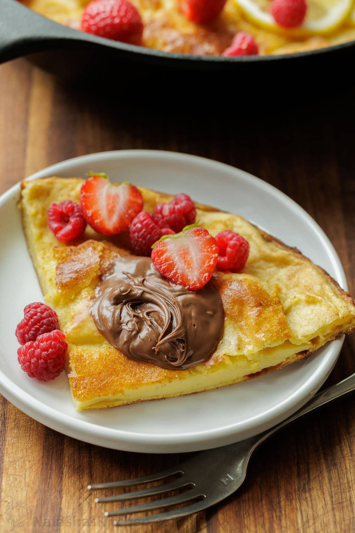 Easy Sourdough Dutch Baby pancake recipe using sourdough discard. Blend or whisk the batter, and it cooks up puffy and golden in the oven. Sourdough Dutch Baby slice on a white plate with Nutella and raspberries on top.