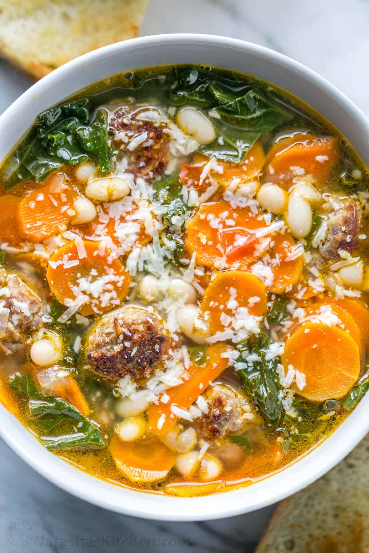 Sausage and Kale Soup is full of veggies and hearty bratwurst sausage cut like meatballs, and it's easy to make in 1 pot and in under 1 hour. White bean, Sausage and kale soup in a white bowl with carrots and parmesan cheese