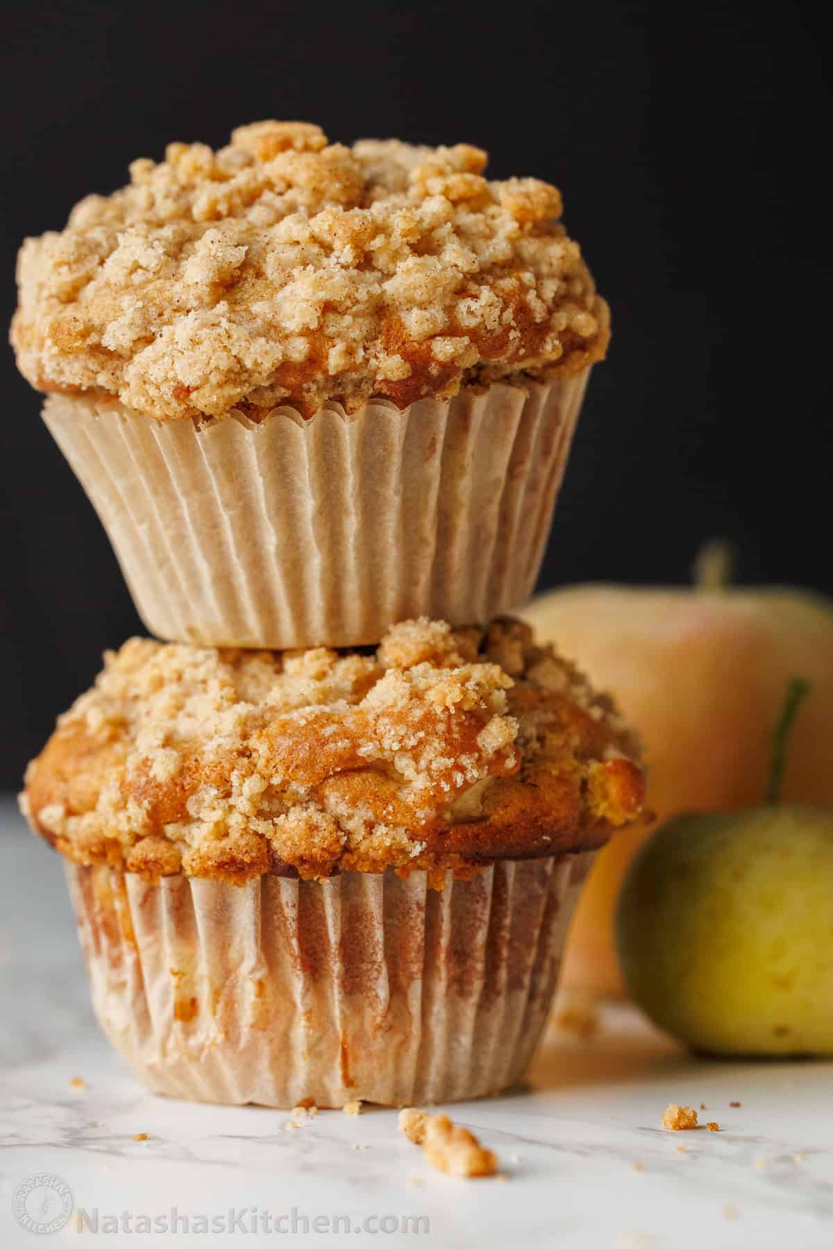 Bakery-style Apple muffins with crumb topping are so easy to make, with a moist crumb and chunks of apple inside. Uses just pantry staples. Two apple muffins stacked on a table with apples in the background