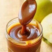 Homemade caramel sauce dripping from a spoon into a mason jar with apples in the background