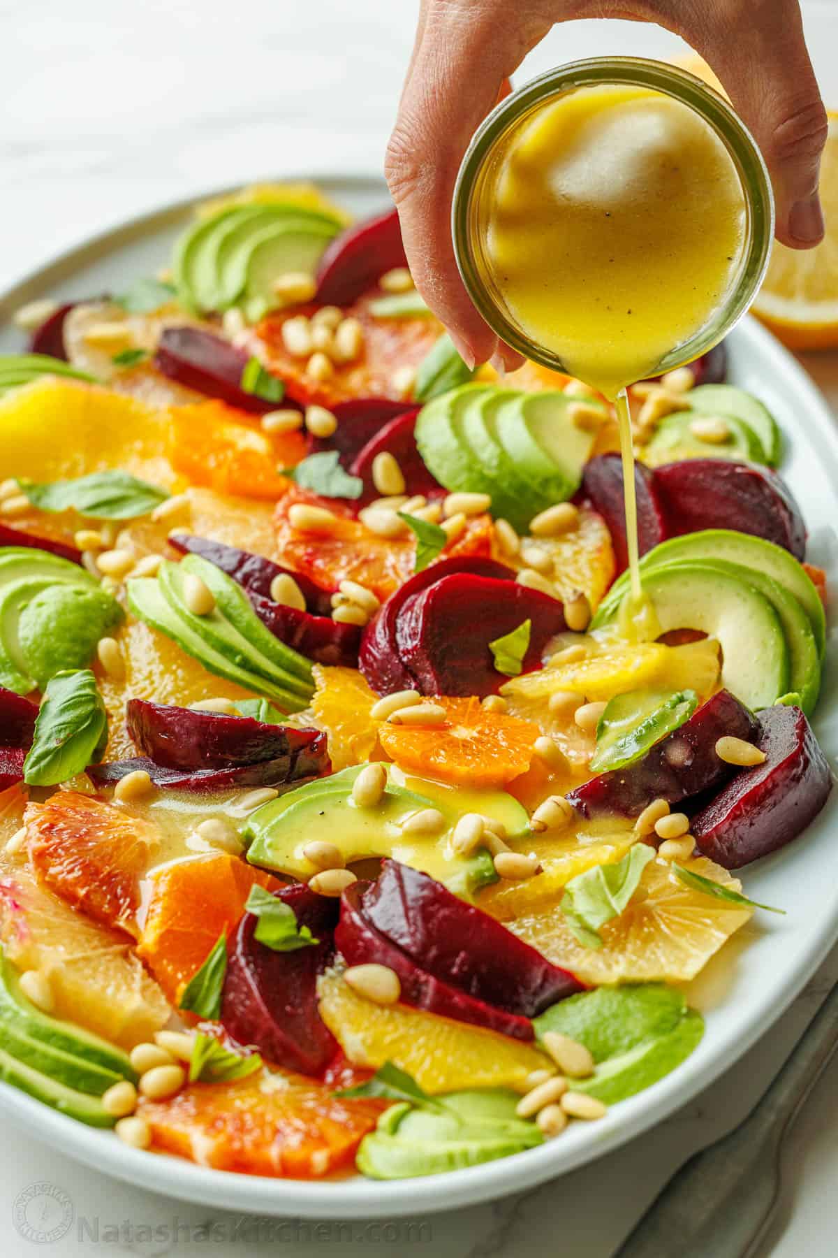 Drizzling lemon vinaigrette over a Citrus salad with fruit and pine nuts on a white platter