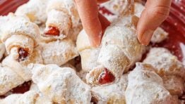 a hand removing one rugelach cookie covered in powdered sugar from a red plate