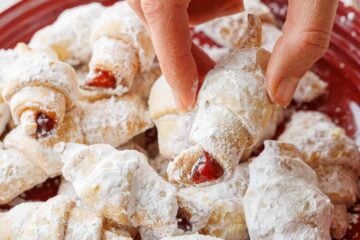 a hand removing one rugelach cookie covered in powdered sugar from a red plate