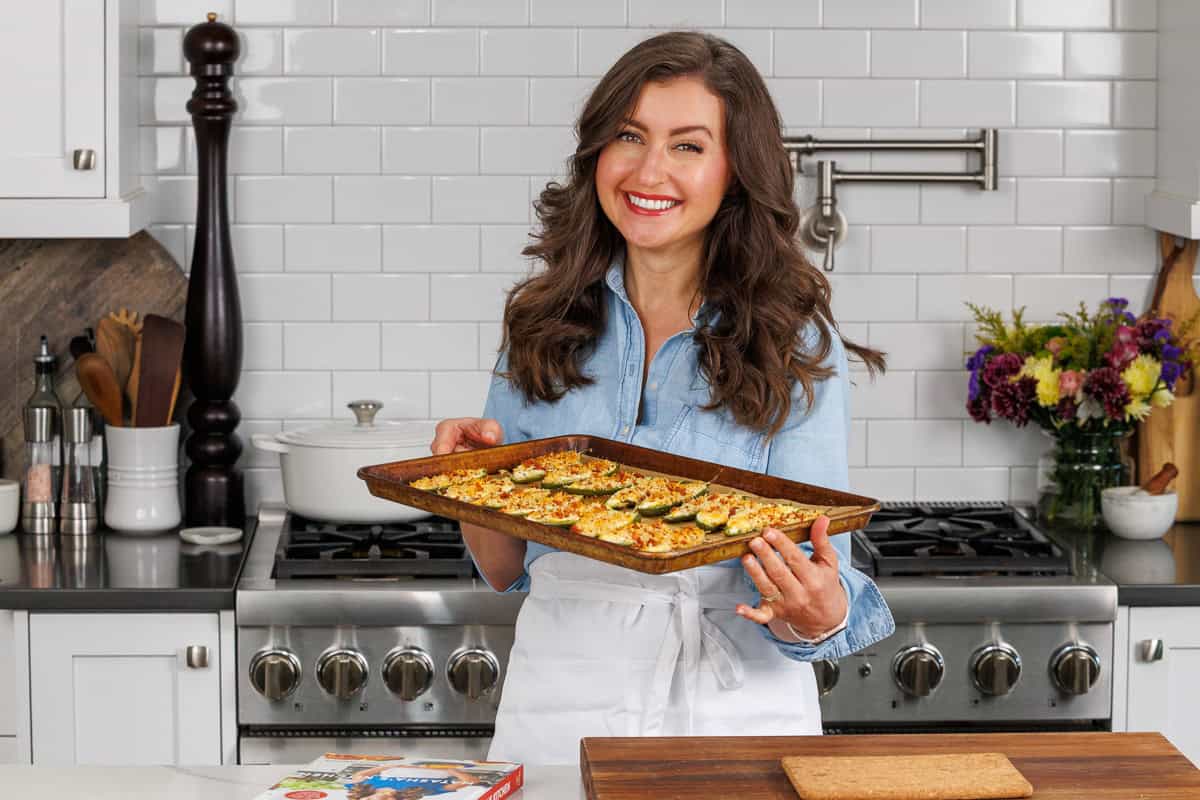 This easy Jalapeno popper recipe is delicious, with a cheesy filling and crisp bacon crumb topping. This crowd-pleasing appetizer goes fast! Natasha holding a tray of jalapeno poppers by the oven