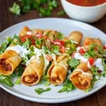easy chicken taquitos recipe on a white plate with salsa for serving and garnished with fresh cilantro, tomatoes, and sour cream drizzle