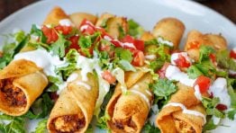 easy chicken taquitos recipe on a white plate with salsa for serving and garnished with fresh cilantro, tomatoes, and sour cream drizzle