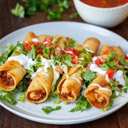 easy chicken taquitos recipe on a white plate with salsa for serving and garnished with fresh cilantro, tomatoes, and sour cream drizzle