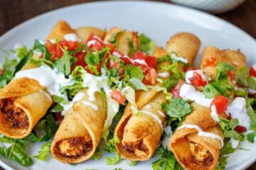 easy chicken taquitos recipe on a white plate with salsa for serving and garnished with fresh cilantro, tomatoes, and sour cream drizzle