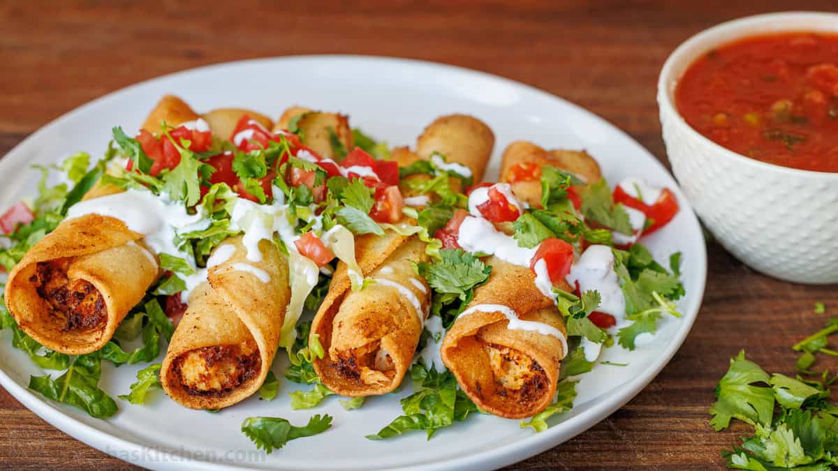 chicken taquitos on a white plate with sour cream, cilantro, and chopped tomato garnish