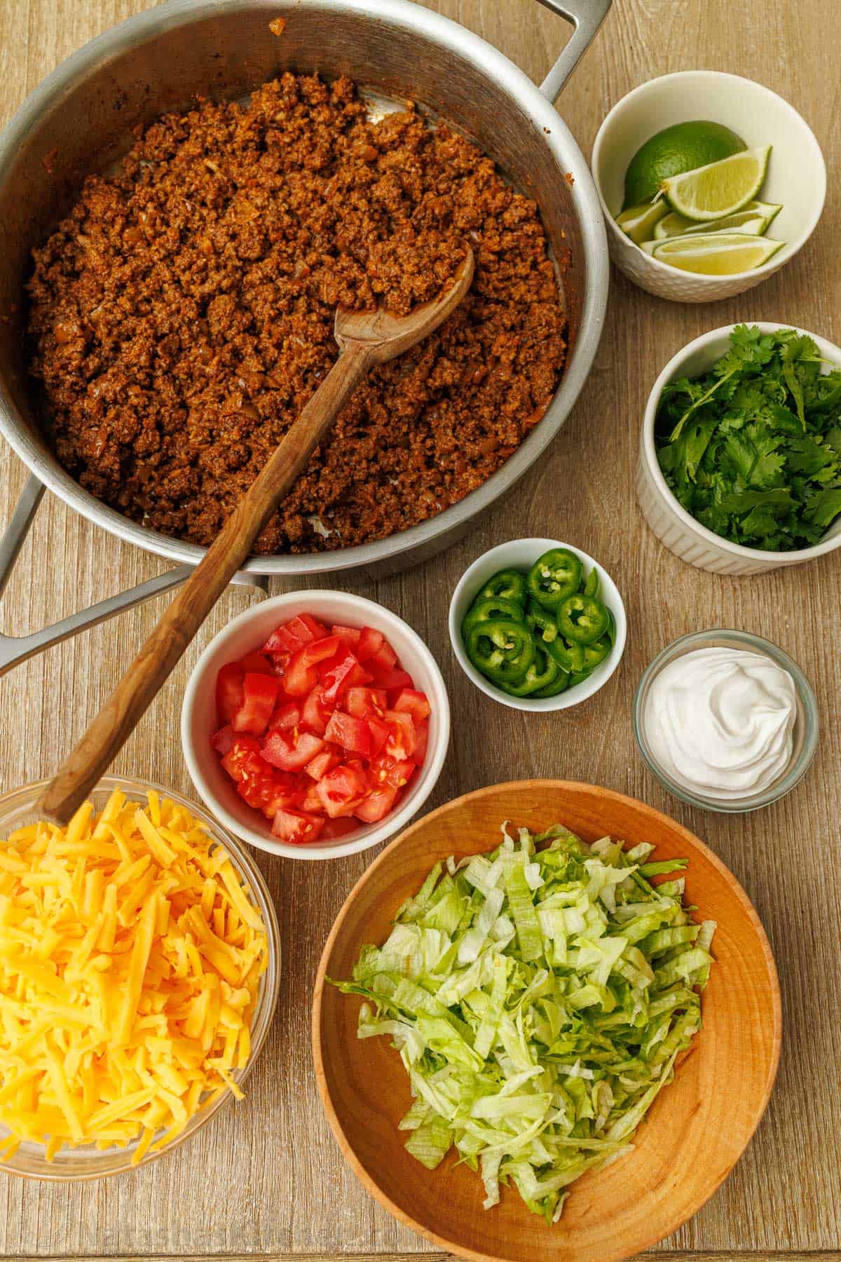 Beef taco meat and taco toppings including lime slices, cilantro, sliced jalapeños, sour cream, chopped tomatoes, shredded cheese and lettuce
