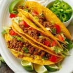 Crunchy ground beef tacos on a white plate with fresh tomatoes, jalapeños, and lime slices as toppings