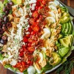 Greek Cobb Salad with ingredients lined up on a plate with tomatoes, bacon, hard boiled eggs, pita chips, avocado, cheese crumbles, chicken and olives
