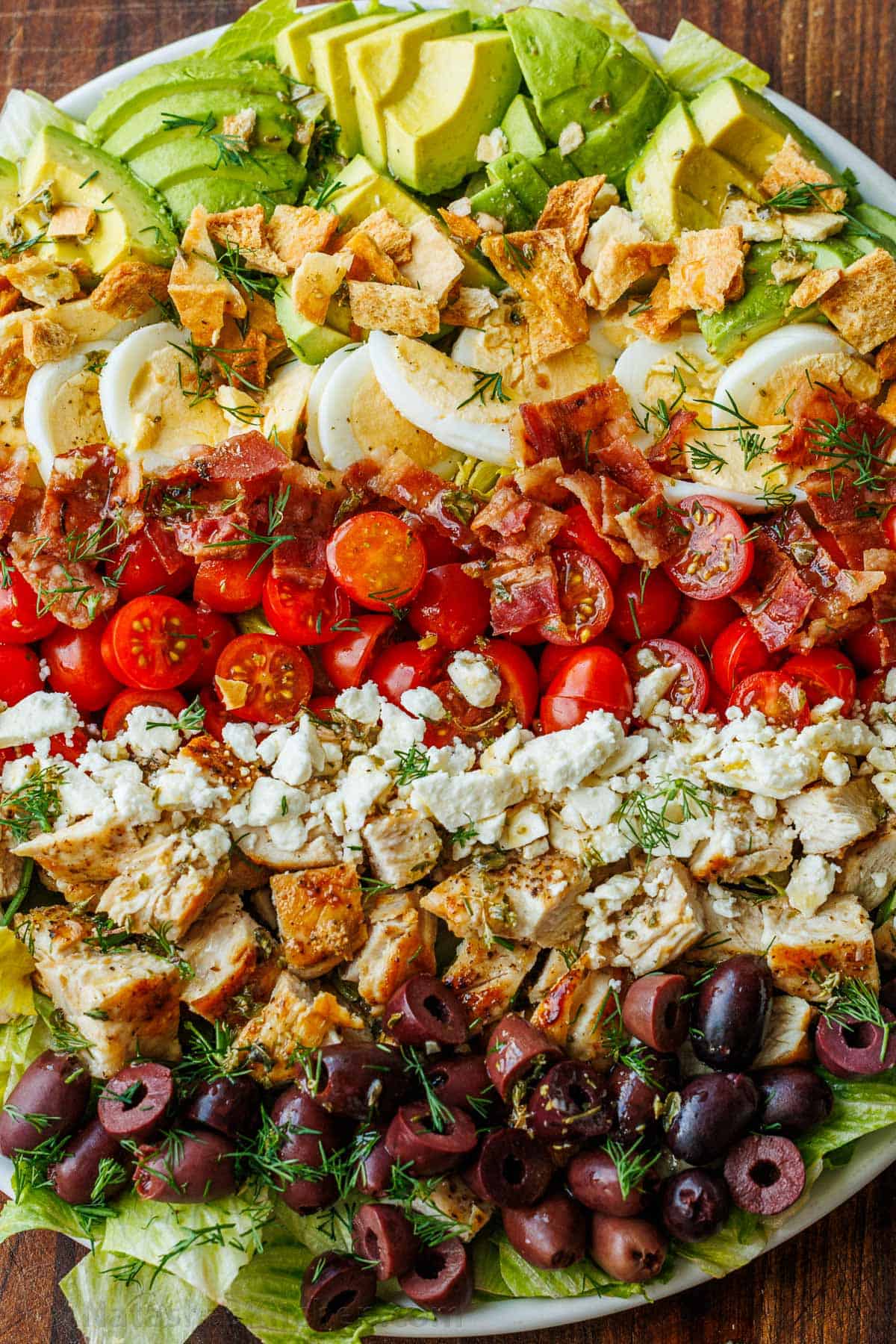 Fresh Greek Cobb salad ingredients lined up on a plate with avocado, pita chips, eggs, tomatoes, bacon, cheese, chicken and olives