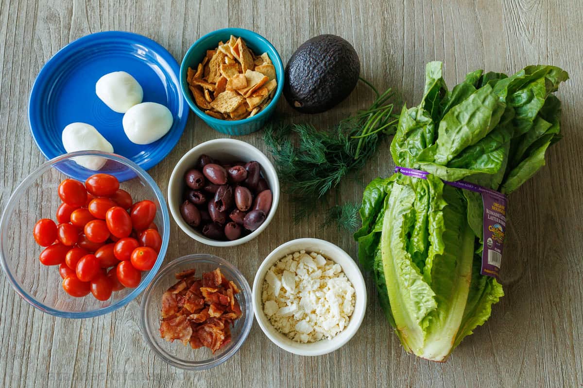 Ingredients for fresh healthy lunch including eggs, tomatoes, olives, bacon, cheese, pita chips, avocado, dill and lettuce