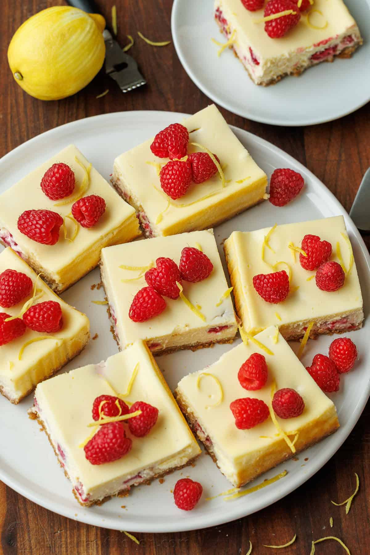 Raspberry lemon cheesecake bars cut in squares on a white plate with raspberries and lemon peel garnish
