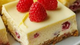 One raspberry lemon cheesecake bar on a plate with raspberries and lemon peel garnish