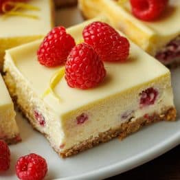 One raspberry lemon cheesecake bar on a plate with raspberries and lemon peel garnish