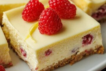 One raspberry lemon cheesecake bar on a plate with raspberries and lemon peel garnish