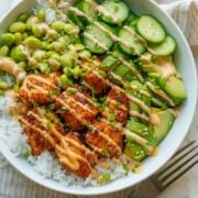 Salmon bowl recipe in a white dish with cucumbers, avocado, seasoned salmon chunks, edamame, rice, and sesame seeds drizzled with 3-ingredient sauce.