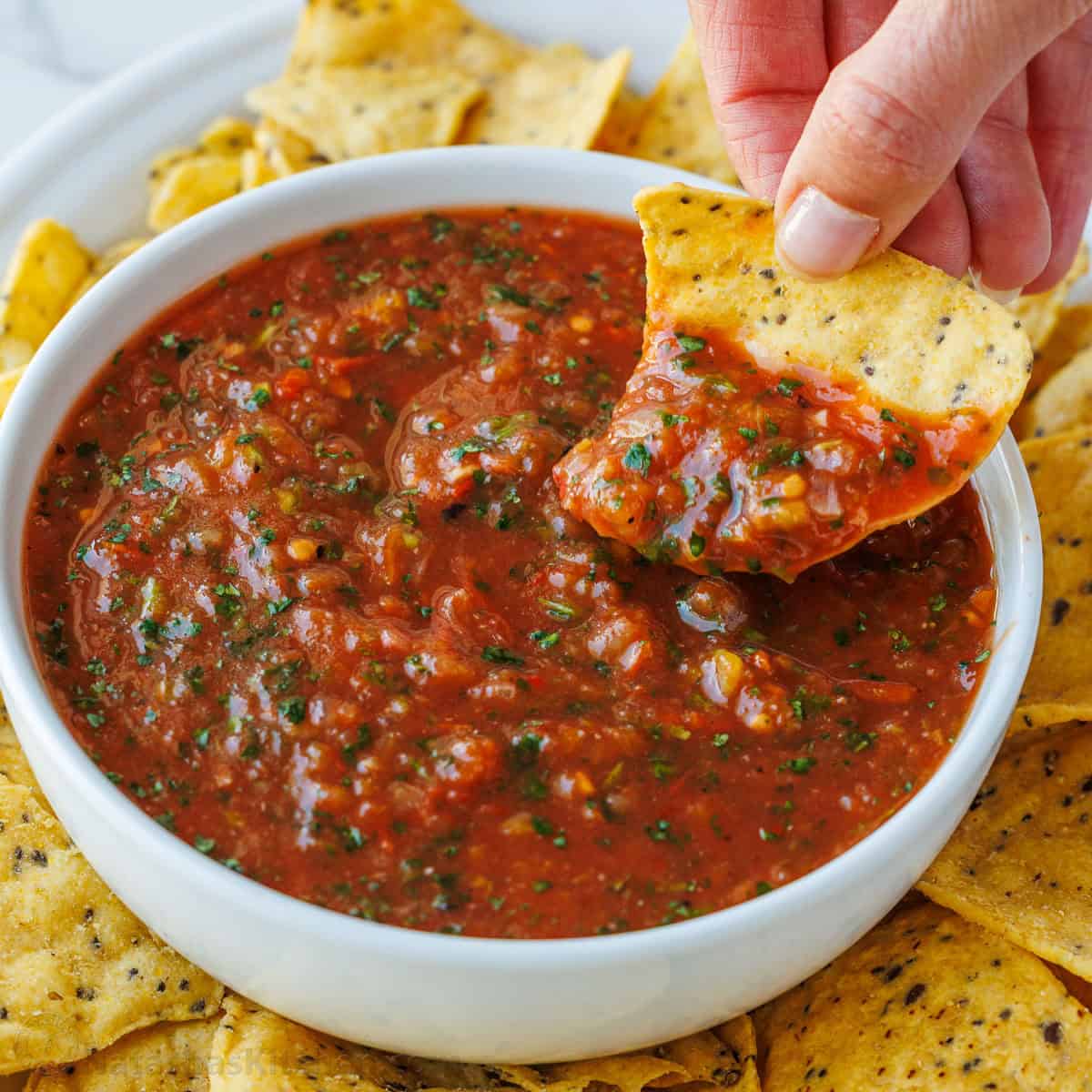 chip scooping roasted tomato salsa from a white bowl
