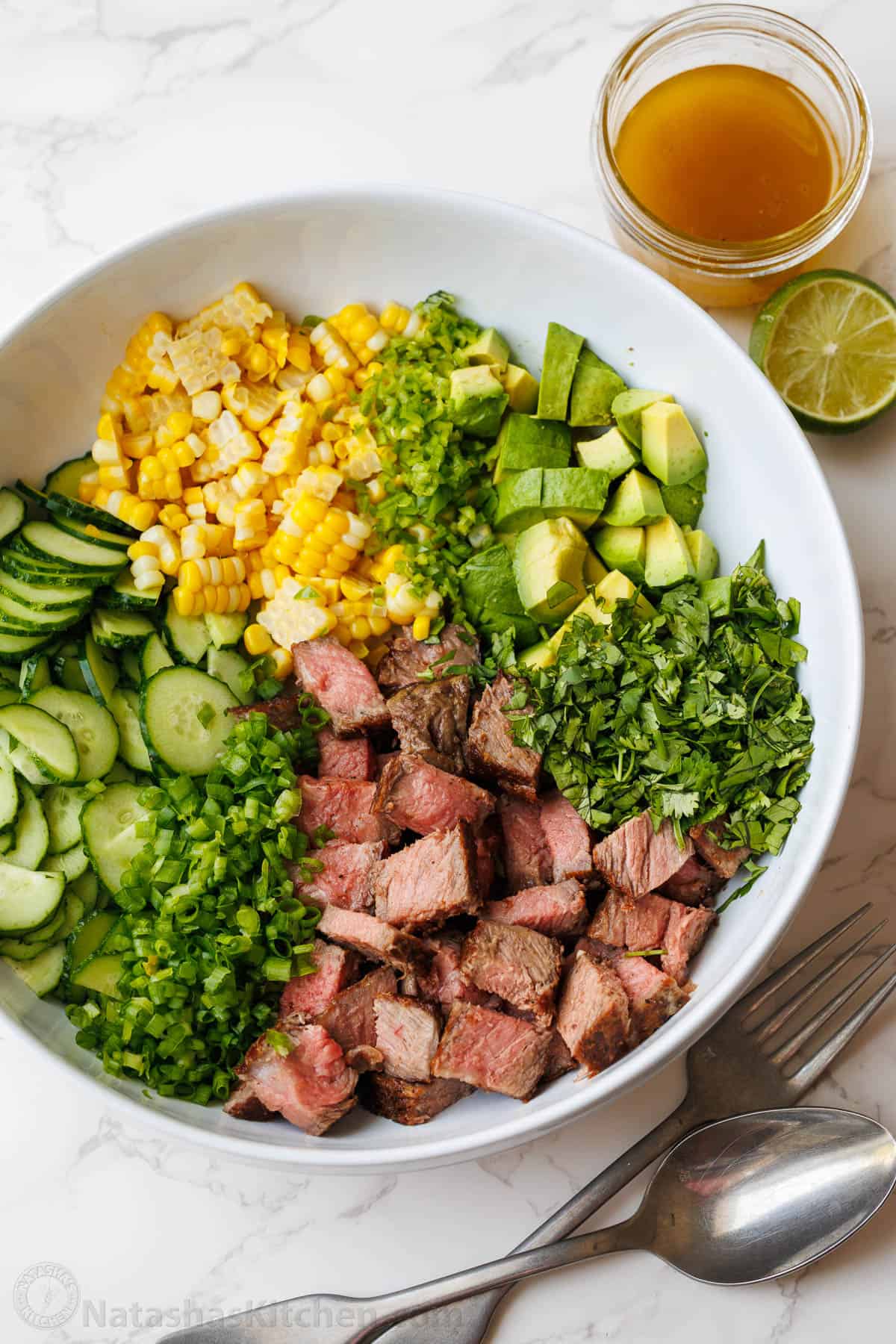 Steak Salad recipe with pan-seared steak, cucumber, corn, avocado, cilantro, and jalapeño, tossed with a vibrant honey-lime dressing. steak salad divided into ingredients in a white bowl with honey lime dressing in a mason jar on the side