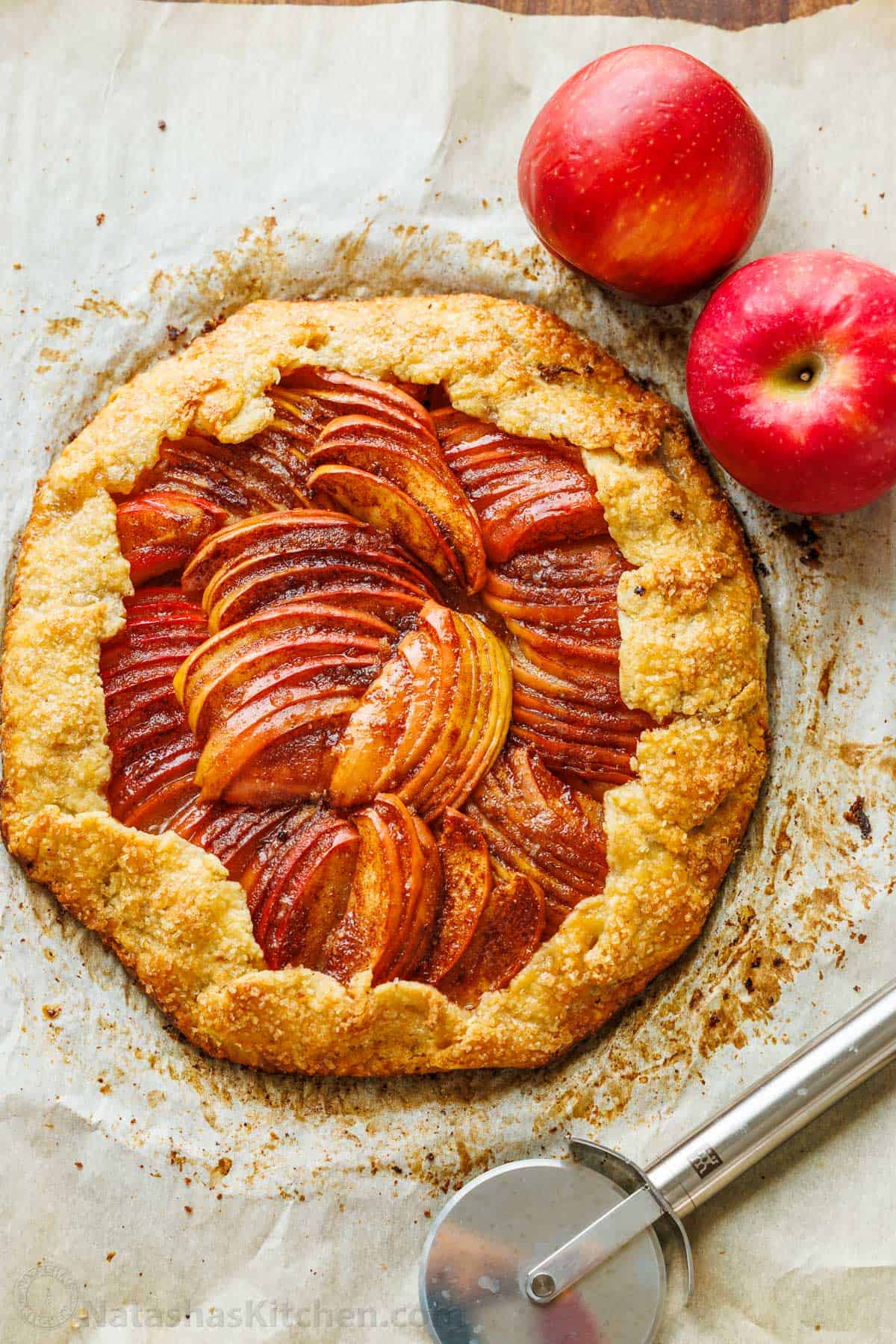 Easy Apple Galette with a flaky homemade crust and cinnamon apples. No pie plate or peeling needed for this beautiful rustic fall dessert. Apple galette on parchment paper with pizza cutter and apples on the side