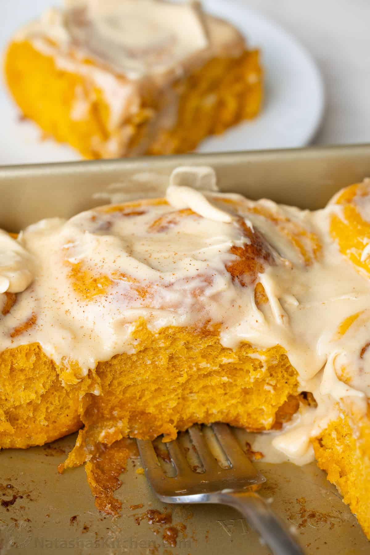 Scooping a pumpkin cinnamon roll out of the pan with frosting on top