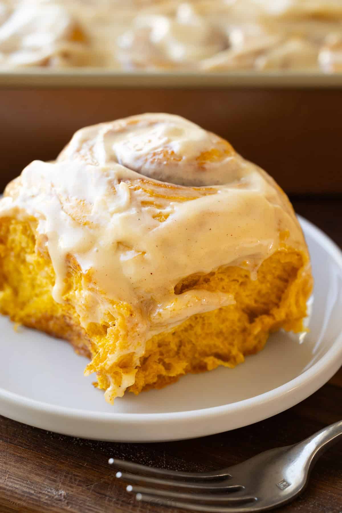 Close up of a pumpkin cinnamon roll on a white plate with icing and a fork to eat with