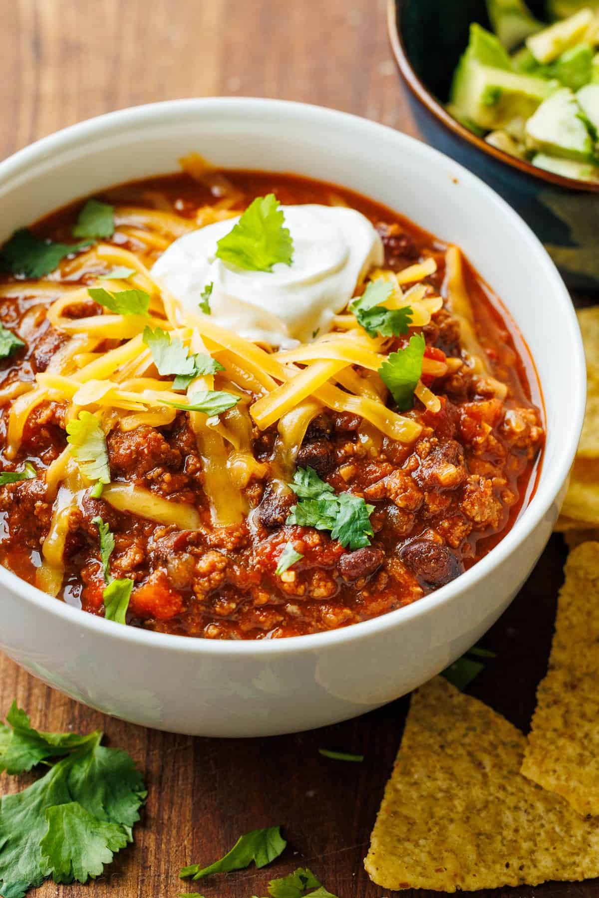 Bowl of ground beef chili topped with cheddar, sour cream and cilantro, with tortilla chips and avocado