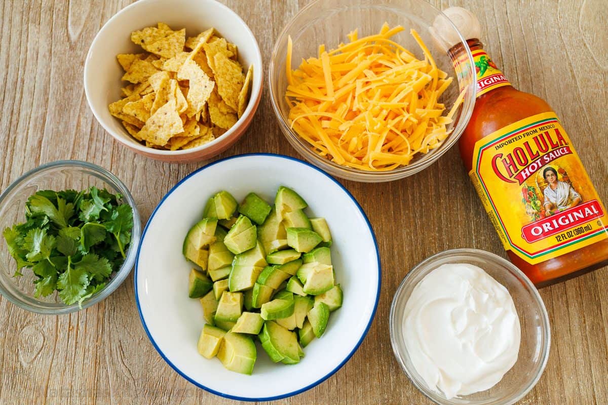 Chili toppings in bowls: tortilla chips, shredded cheddar, cilantro, diced avocado, sour cream and a bottle of hot sauce