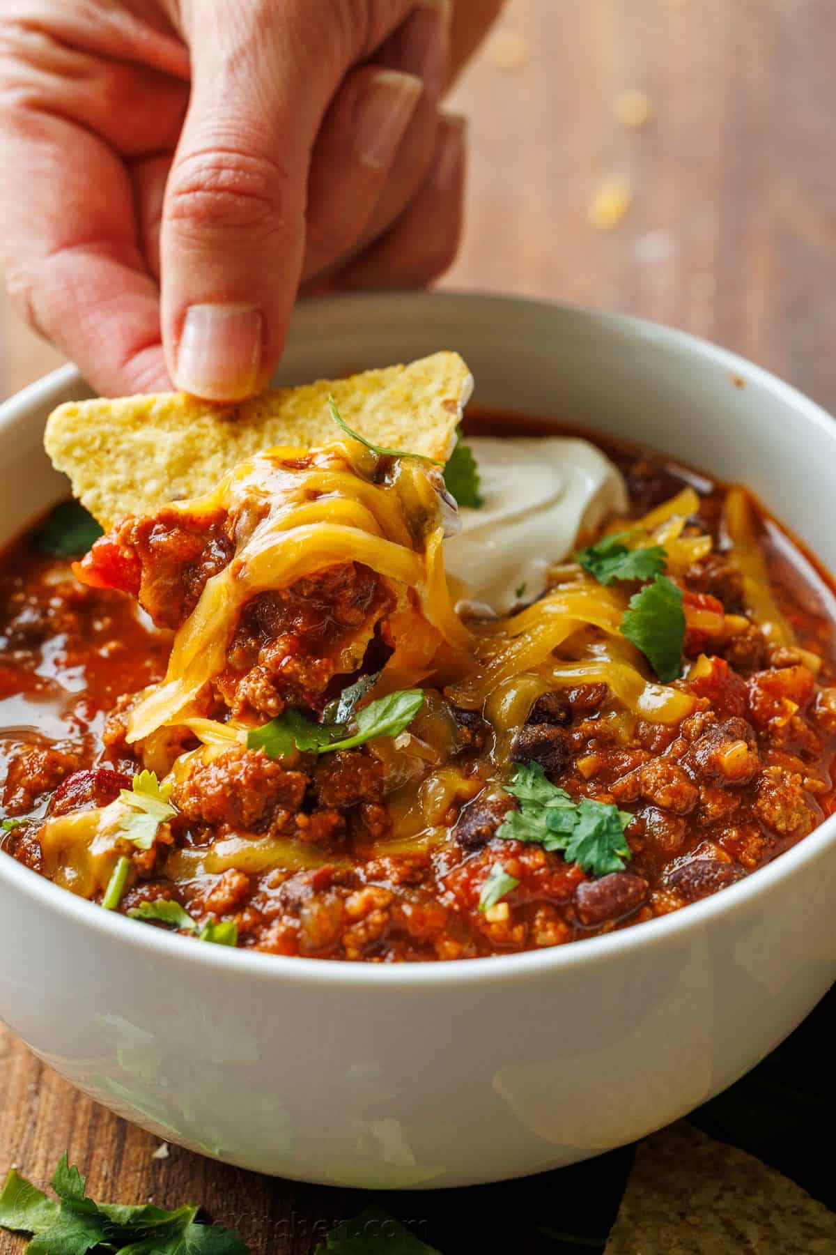 Hand dipping a tortilla chip into a bowl of beef chili with melted cheddar and sour cream