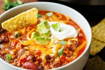 Bowl of Instant Pot chili topped with shredded cheddar, sour cream, avocado, cilantro and a tortilla chip in front of an Instant Pot